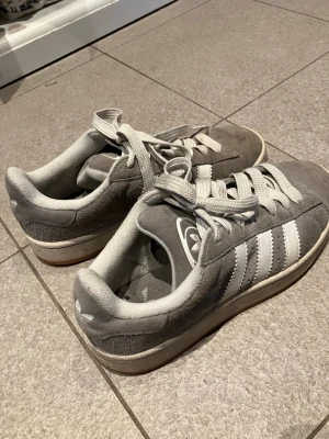 Grå Adidas Campus 38 2/3 - Säljer ett par grå Adidas Campus sneakers med klassiska vita ränder på sidan. Skorna har rund tå, platt sula och är tillverkade i mocka med vita detaljer och snörning. Perfekta för dig som gillar retrostil och vill ha något bekvämt till vardags.