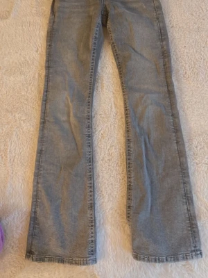 Bootcut ljusgrå jeans från Gina  - Säljer ett par ljusgrå bootcut jeans med snygga fickdetaljer bak, inklusive dekorativa knappar och mönstrad. Passformen är normal och benen är lätt utsvängda nedtill.