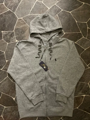 Grå hoodie från Polo Ralph Lauren - Snygg grå hoodie från Polo Ralph Lauren med dragkedja och huva. Tröjan har snörning vid halsen, ribbade muddar och klassisk Polo-logga broderad på bröstet. Perfekt för chill dagar och enkel att matcha med jeans eller joggers. Plagget sitter som M - S, något mellan där.