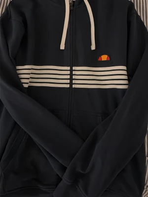 Svart kofta från Ellesse - Säljer en svart hoodie från Ellesse i storlek L. Tröjan har vita snören, dragkedja och vita ränder över bröstet. Notera att passformen är M!!! 