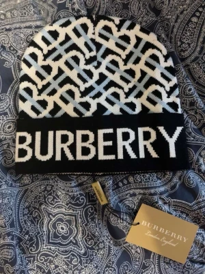 Burberry mössa med logomönster - Säljer en snygg Burberry mössa med stort logomönster i svart, vitt och ljusblått. Mössan har bred svart kant med BURBERRY-text framtill och är stickad i mjukt material. Perfekt för dig som vill ha en statement-accessoar med lyxig känsla.