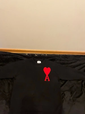 Svart stickad tröja från Ami Paris - Svart stickad tröja från Ami Paris i storlek S. Tröjan har ett stort rött hjärta och bokstaven A broderat på bröstet. Rund hals och långa ärmar, perfekt för dig som gillar stilrena detaljer med en twist.