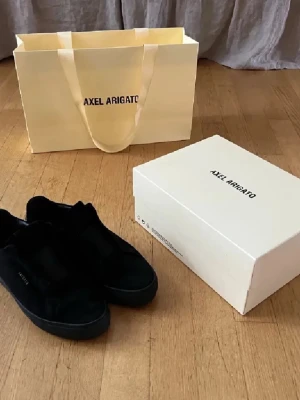 Svarta sneakers från Axel Arigato - Stilrena svarta sneakers från Axel Arigato i mocka med diskret logga i guld på sidan. Skorna har en klassisk låg profil och platt sula, perfekt för en clean och modern look. Kommer med originalkartong och påse.