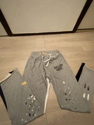 Grå mjukisbyxor från Gallery Dept. - Säljer ett par grå mjukisbyxor från Gallery Dept. med vit snörning i midjan och coola färgstänk i svart, vitt och gult på benen. Byxorna har tryckt logga på framsidan och vita detaljer längs sidorna. Perfekt för dig som gillar streetwear och unik design.