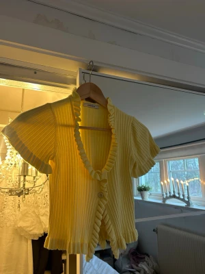 Gul ribbad kofta från Chiquelle - Säljer en söt gul ribbad kofta från Chiquelle med korta ärmar och volangkant längs v-ringningen och nederkanten. Koftan har två knappar framtill och är perfekt för vår och sommar. Superfin att styla med jeans eller kjol!