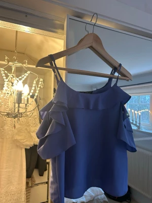 Blå volangblus med smala axelband - Säljer en blå blus med smala axelband och volangdetaljer längs axlarna och halsringningen.