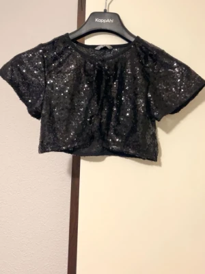 Svart paljett bolerotopp  - Säljer en svart bolerotopp, täckt med glittriga paljetter. Toppen är croppad med korta ärmar och rund halsringning. Perfekt för att addera lite extra bling till din outfit. Snygg och trendig design som sticker ut. Passar xxs,xs,164 barn