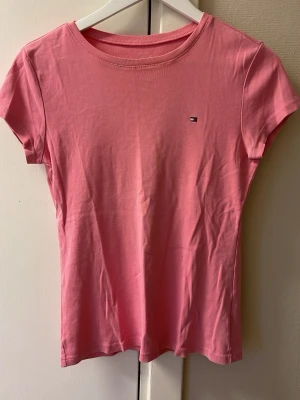 Rosa t-shirt från Tommy Hilfiger - Snygg rosa t-shirt från Tommy Hilfiger med liten broderad logga på bröstet. Modellen är klassisk med rund hals och korta ärmar, perfekt för en chill och enkel stil. Mjuk bomull som känns skön mot huden och passar till jeans eller shorts.
