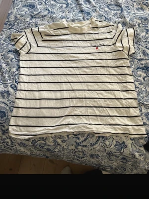 Vit randig t-shirt från H&M - Säljer en vit t-shirt från H&M med svarta horisontella ränder och broderad text 'Livres et Liberté' samt ett litet rött hjärta på bröstet. T-shirten har rund hals och korta ärmar, tillverkad i mjuk bomull. Perfekt för chill dagar och enkel att matcha.