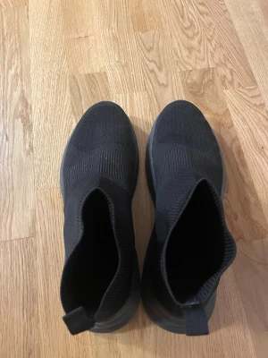 Svarta stickade sneakers med platt sula.   - Säljer ett par helsvarta sneakers med sockliknande design och platt sula. Skorna är stickade i syntetmaterial och har en minimalistisk look utan snörning. Perfekta för dig som gillar en clean och modern stil. Helt oanvända 