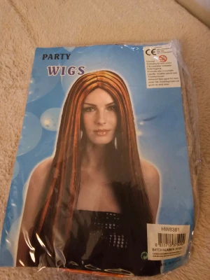 Svart och orange peruk med långa slingor - Cool peruk med långa, raka slingor i svart och orange. Perfekt för att sticka ut på festen eller maskerad! Syntetmaterial som är lätt att styla och använda. Säljs i originalförpackning.