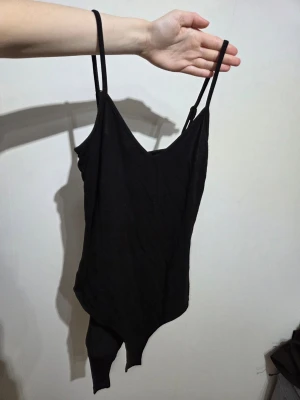 Svart bodysuit från H&M i XS - Säljer en svart bodysuit från H&M i storlek XS. Den har smala axelband, v-ringad hals och tryckknappar i grenen. Materialet är mjukt och stretchigt, troligen bomull med inslag av syntet. Perfekt att styla med jeans eller kjol.