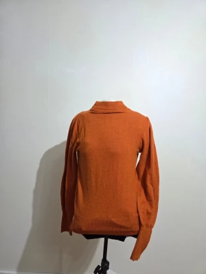 Rostorange polotröja från KappAhl - Snygg rostorange polotröja från KappAhl i storlek S. Tröjan har puffade axlar och långa ärmar med ribbade muddar. Materialet är mjukt och stickat, perfekt för kyliga dagar. Polokragen är ribbad och ger en cool touch till looken.