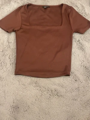 Brun topp med fyrkantig halsringning - Säljer en enkel brun topp med korta ärmar och snygg fyrkantig halsringning. Toppen har en slim passform och är tillverkad i mjukt bomullsmaterial. Perfekt att styla med jeans eller kjol för en clean look. Kan eventuellt tänka mig gå ner i pris