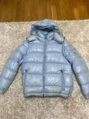 Ljusblå Moncler Maya - Säljer en ljusblå dunjacka från Moncler med huva och dragkedja framtill. Jackan har två fickor med dragkedja och ett Moncler-märke på ärmen. Den är quiltad och har en snygg, glansig finish som gör den extra stilren för vintern. Använd max 10 gånger och den är Riktig