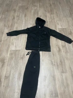 Svarta mjukisbyxor från Lacoste - Säljer ett set svarta mjukisbyxor och kofta från Lacoste med dragsko i midjan och muddar vid bensluten. Byxorna har den klassiska gröna krokodilloggan broderad på benet och är tillverkade i mjuk bomull för maximal komfort. Koftan har kön bomull material för maximal komfort