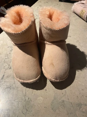 Ljus rosa UGG boots med fluffig insida - Säljer ett par ljus rosa UGG boots för barn med mjuk och fluffig insida. Skorna är tillverkade i mocka och har rund tå samt platt sula. Klassisk UGG-logga på sidan och grova sömmar runt kanterna. Perfekta för kalla dagar nu i vinter.
