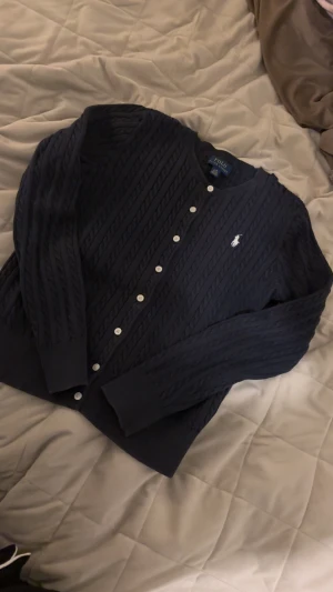 Mörkblå kofta från Polo Ralph Lauren - Snygg mörkblå kofta från Polo Ralph Lauren med kabelstickat mönster och vita knappar framtill. Klassisk rund hals och broderad logga på bröstet. Perfekt för lager-på-lager och chill stil. Materialet är mjukt och stickat för extra komfort.