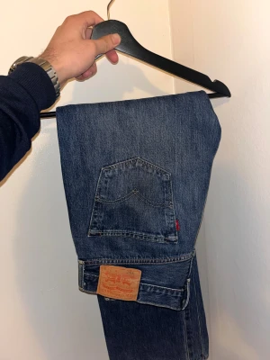 Levi's 501 blå jeans - Klassiska Levi's 501 jeans i blå denim med raka ben och normal passform. Snygga kontrastsömmar och ikonisk läderpatch bak i midjan. Jeansen har fem fickor och den typiska röda Levi's-taggen på bakfickan. Perfekt för dig som gillar tidlös stil. W33 L30