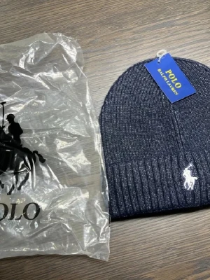 Mörkblå ribbad mössa från Polo Ralph Lauren - Snygg mörkblå ribbstickad mössa från Polo Ralph Lauren med klassisk vit logga framtill. Mössan har uppvikt kant och är tillverkad i mjukt material som håller dig varm. Perfekt accessoar för kalla dagar och enkel att matcha med din stil.