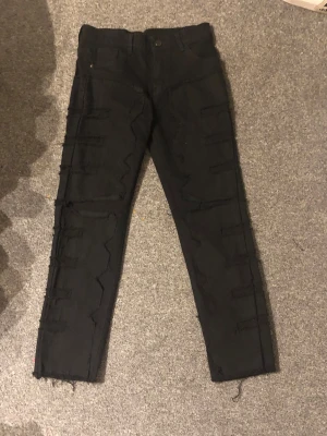 Svarta jeans med råa detaljer - Säljer ett par svarta jeans med råa, fransiga detaljer längs benen och en smal passform. Byxorna har klassisk femficksdesign och knappstängning. Materialet är jeans och färgen är helsvart, vilket ger en cool och edgy vibe.
