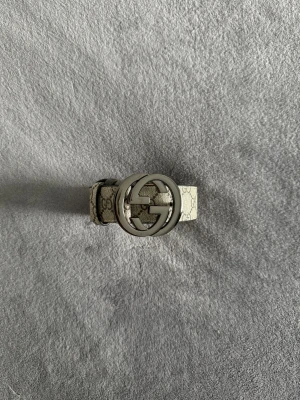Gucci bälte  - Säljer ett snyggt Gucci-skärp i beige med klassiskt GG-mönster och stor silverfärgad logospänne. Skärpet är gjort i skinn och har en lyxig känsla med detaljerad söm på insidan. Perfekt för att lyfta din outfit med en ikonisk accessoar.