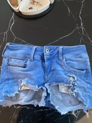 Blå jeansshorts med slitningar - Säljer ett par blå jeansshorts med råa, slitna kanter och hål för en edgy look. Shortsen har klassisk femficksdesign, knapp och dragkedja framtill samt bälteshällor. Perfekta för dig som gillar streetstyle och vill sticka ut.