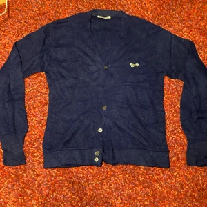 Blå kofta från the fox med räv - Snygg marinblå kofta från JC Penney med broderad grön räv på bröstet. Koftan har v-ringad hals, långa ärmar och knappar framtill. Materialet är stickat och känns mjukt mot huden. Perfekt för dig som gillar klassisk stil med en twist.