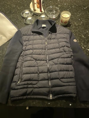 Svart quiltad pufferjacka från Moncler - Säljer en navy pufferjacka från Moncler med quiltad front och släta stickade ärmar. Jackan har två dragkedjefickor, hel dragkedja och klassisk Moncler-logga på ärmen. Perfekt för dig som vill ha en snygg och varm jacka med exklusiv känsla. Jackan passar allt från L/M