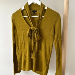 Senapsgul tröja med knyt från Massimo Dutti - Senapsgul långärmad tröja från Massimo Dutti med v-ringning och långa band som kan knytas i en rosett framtill. Tunn och mjuk i bomullsmix. Snygg detalj med knyt.