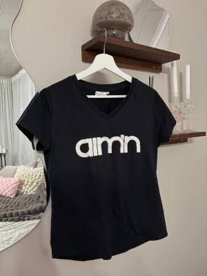 Svart t-shirt med v-ringning från amn - Svart t-shirt med vit amn-logga på bröstet. T-shirten har v-ringad hals och korta ärmar, perfekt för en chill och stilren look. Materialet känns mjukt och skönt mot huden, och passformen är normal. Enkelt att matcha med jeans eller shorts.