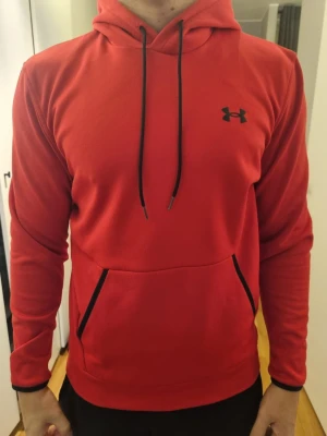 Röd hoodie från Under Armour - Snygg röd hoodie från Under Armour med svart logga på bröstet. Tröjan har huva med svarta snören och en stor magficka framtill. Inga byten.