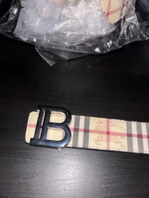 Beige rutig väst med B-spänne - Säljer en beige väst med klassiskt rutigt mönster i svart, rött och grått. Västen har ett stort metallspänne med bokstaven B framtill. Materialet är skinn och designen är riktigt stilren och modern, perfekt för dig som vill sticka ut med en ikonisk detalj. OBS INTE ORG1NAL