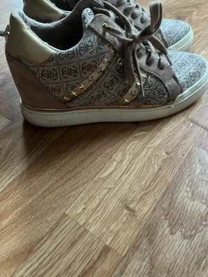 Beige sneakers från Guess med mönster - Säljer ett par beige sneakers från Guess med logomönster över hela skon och guldfärgade detaljer med stjärnor på sidan. Skorna har snörning, rund tå och platt sula. Materialet är en mix av textil och mocka med inslag av syntet vid hälen.