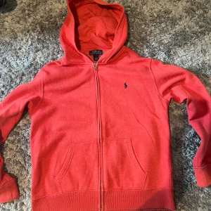 Röd hoodie från Polo Ralph Lauren - Säljer en röd hoodie från Polo Ralph Lauren med dragkedja och huva. Tröjan har två fickor framtill och den klassiska broderade loggan på bröstet. Perfekt för dig som gillar en avslappnad och sportig stil. Storlek 14-16 år (L) barnstorlek. Väldigt fint skick.