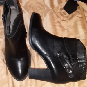 Svarta stövletter med klack och spänne tamaris - Snygga svarta stövletter från tamaris med markerad klack och rund tå. Skorna är gjorda i skinn och har dekorativa remmar med metallknappar och spänne runt ankeln. Perfekt för dig som gillar stilrena boots med lite edge. Använda 1 gång 