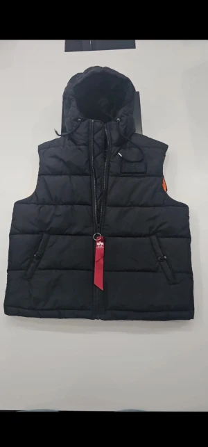 Svart dunväst Alpha Industries M - Säljer en svart dunväst från Alpha Industries med huva och orange innerfoder. Västen har två fickor med knappar, dragkedja framtill och en röd tag med logga. Perfekt för lager-på-lager och streetwear-vibe.