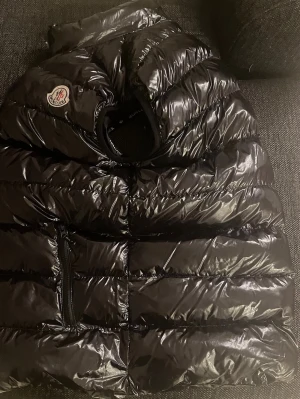 Svart dunväst från Moncler - Säljer en riktigt fet, svart, glansig dunväst från Moncler med klassisk logga på bröstet. Västen är i väldigt bra skick och inte använd så många gånger. Perfekt för dig som vill ha en snygg och varm look.