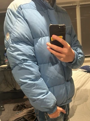 Ljusblå Moncler pufferjacka - Säljer en ljusblå pufferjacka från Moncler med klassisk logga på ärmen. Jackan har hög krage, långa ärmar och stängs med dragkedja och knappar. Fodret är svart och materialet är polyester. Perfekt för kalla dagar och riktigt snygg till jeans. 