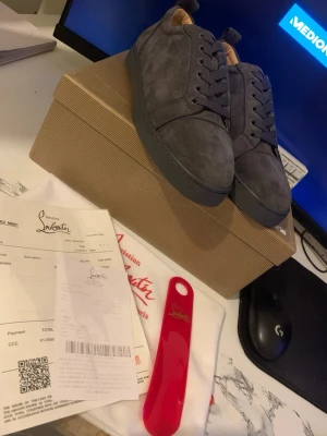 Grå mocka sneakers från Louboutin - Snygga grå sneakers från Christian Louboutin i mjuk mocka med klassisk röd sula och rund tå. Skorna har snörning och en stilren design som passar perfekt till streetwear. Kommer med originalkartong, dustbag och skohorn.