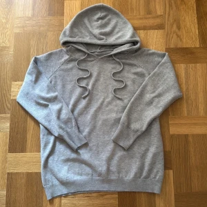 Grå kashmir hoodie - En enkel och stilren grå kashmir hoodie med huva och snörning framtill. Tröjan har långa ärmar och breda ribbade muddar vid ärmslut och nederkant. Perfekt för chill dagar och passar till det mesta. Materialet känns mjukt och är väldigt skönt enligt mig.