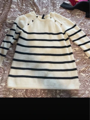 Randig stickad tröja  - Säljer en vit och svart randig stickad tröja från i storlek L. Tröjan har långa ärmar, ribbade muddar och dekorativa knappar vid axlarna. Perfekt för dig som gillar klassisk stil med en twist. Osäker på märket men har knappt använt denna tröjan. 