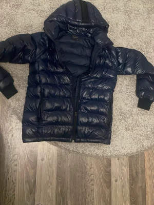 Blå Peak Performance pufferjacka - Säljer en mörkblå pufferjacka från Peak Performance med huva och svarta muddar. Jackan har dragkedja framtill, två sidofickor med dragkedja och Peak Performance-logga på ärmen. Perfekt för kalla dagar och riktigt snygg streetstil.