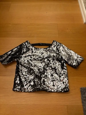 Glittrig paljettopp från Bik Bok, storlek XS - Säljer en glittrig topp från Bik Bok i storlek XS. Toppen är täckt av svarta och silvriga paljetter, har korta ärmar och en snygg öppen rygg. Perfekt för dig som vill sticka ut med en festlig look! 100% Polyester.
