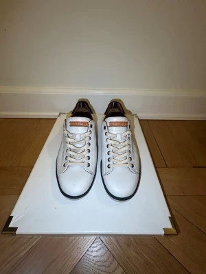 Tommy Hilfiger vita sneakers✨ - Oandvända snygga vita sneakers från Tommy Hilfiger med klassisk design och bruna detaljer på insidan. Skorna har snörning, rund tå och svart kant vid sulan. Materialet är skinn och insidan är i brunt läder. Perfekt för dig som gillar stilrena och grisch sneakers.