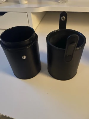 Svart cylinderformad sminkborstfodral - Snyggt svart cylinderformat fodral för sminkborstar i konstläder. Stängs med tryckknappar på sidorna och kan delas i två delar. Perfekt för att hålla borstar organiserade både hemma och på resan. Mått: 23,5 x 8,8 x 10,3 cm.