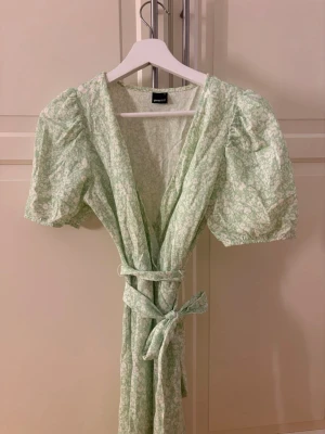 Grön blommig omlottklänning Gina Tricot - Supersöt ljusgrön omlottklänning från Gina Tricot med puffärmar och knytband i midjan. Klänningen har ett vitt blommönster och v-ringad urringning. Perfekt för sommarens alla tillfällen!