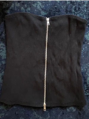 Svart ribbad tubtopp med dragkedja - Säljer en svart ribbad tubtopp från Weekday med en snygg silverfärgad dragkedja framtill. Toppen är axelbandslös och har en croppad passform som ger en cool vibe. 