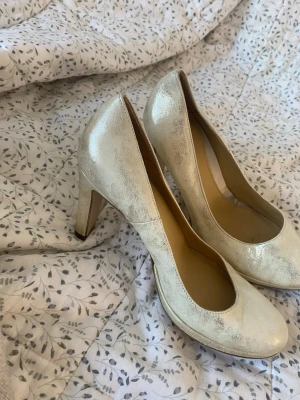 Guldiga pumps från Dasia, storlek 38 - Säljer ett par eleganta pumps från Dasia i storlek 38. Skorna är i skimrande guldigt skinn med rund tå och hög klack. 