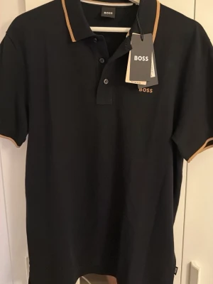 Svart piké från BOSS med beige detaljer - Snygg svart pikétröja från BOSS med beige kantband på krage och ärmslut.  Storleken är M och den e helt ny, inte använd en ända gång 